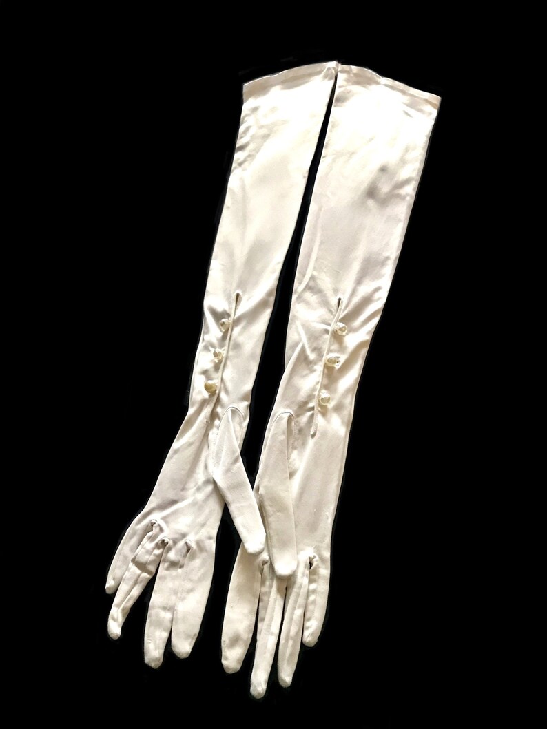 May include: A pair of long white satin gloves with pearl buttons. The gloves are fully extended and the fingers are spread out.