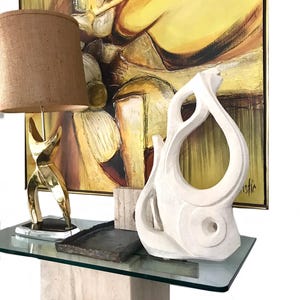 Pode incluir: Um candeeiro de mesa em lat&atilde;o com c&uacute;pula de juta castanha, um tabuleiro retangular escuro e uma escultura abstrata branca com recortes org&acirc;nicos. Estes itens est&atilde;o dispostos numa mesa de consola de vidro em frente a uma pintura.