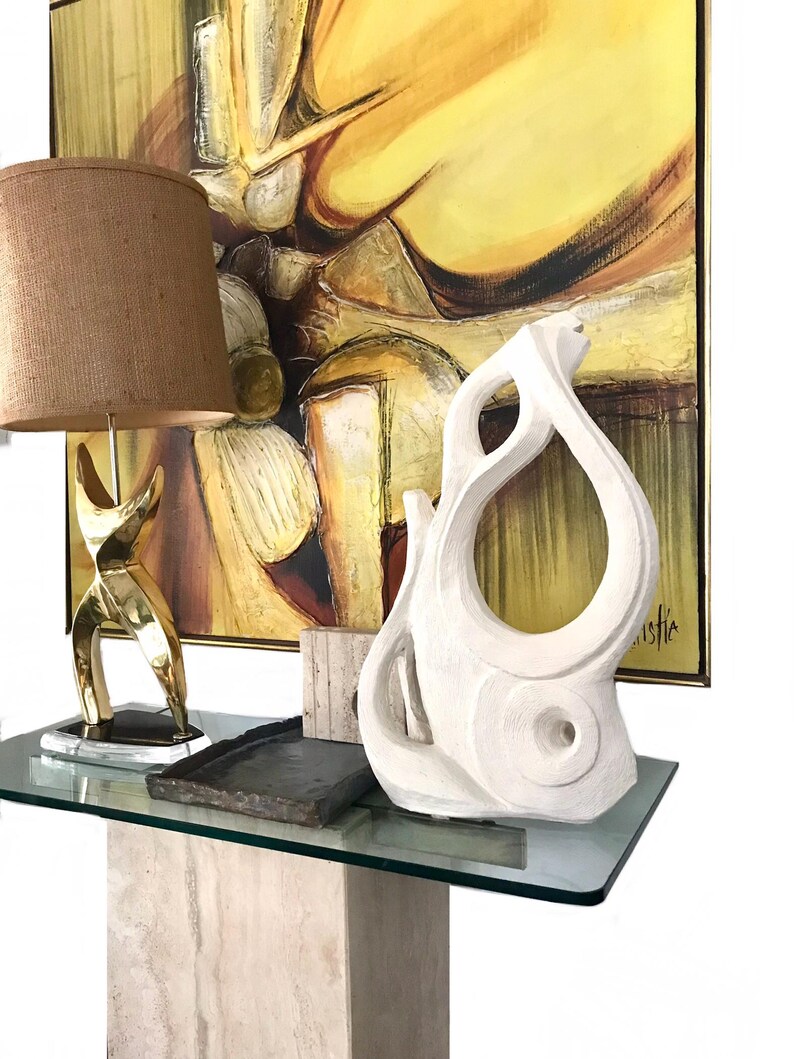 Pode incluir: Um candeeiro de mesa em lat&atilde;o com c&uacute;pula de juta castanha, um tabuleiro retangular escuro e uma escultura abstrata branca com recortes org&acirc;nicos. Estes itens est&atilde;o dispostos numa mesa de consola de vidro em frente a uma pintura.
