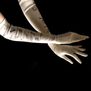 May include: A pair of long, white satin gloves with a whimsical design of keys, tents, and other symbols. The gloves are worn on a person's arms, with the hands extended.