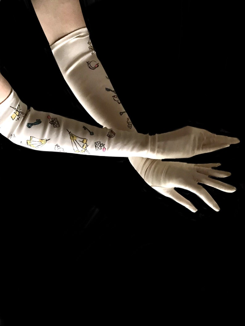 May include: A pair of long, white satin gloves with a whimsical design of keys, tents, and other symbols. The gloves are worn on a person's arms, with the hands extended.