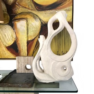 Pode incluir: Uma escultura abstrata branca e texturizada com recortes org&acirc;nicos curvos e uma abertura circular. Ao lado, h&aacute; um pequeno bloco de pedra bege quadrado com um furo e uma bandeja de pedra escura, retangular e de bordas rugosas.