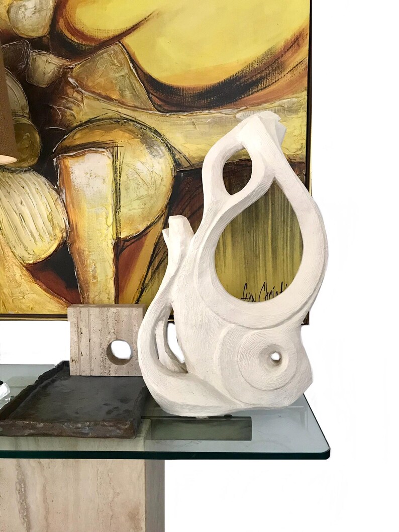 Pode incluir: Uma escultura abstrata branca e texturizada com recortes org&acirc;nicos curvos e uma abertura circular. Ao lado, h&aacute; um pequeno bloco de pedra bege quadrado com um furo e uma bandeja de pedra escura, retangular e de bordas rugosas.