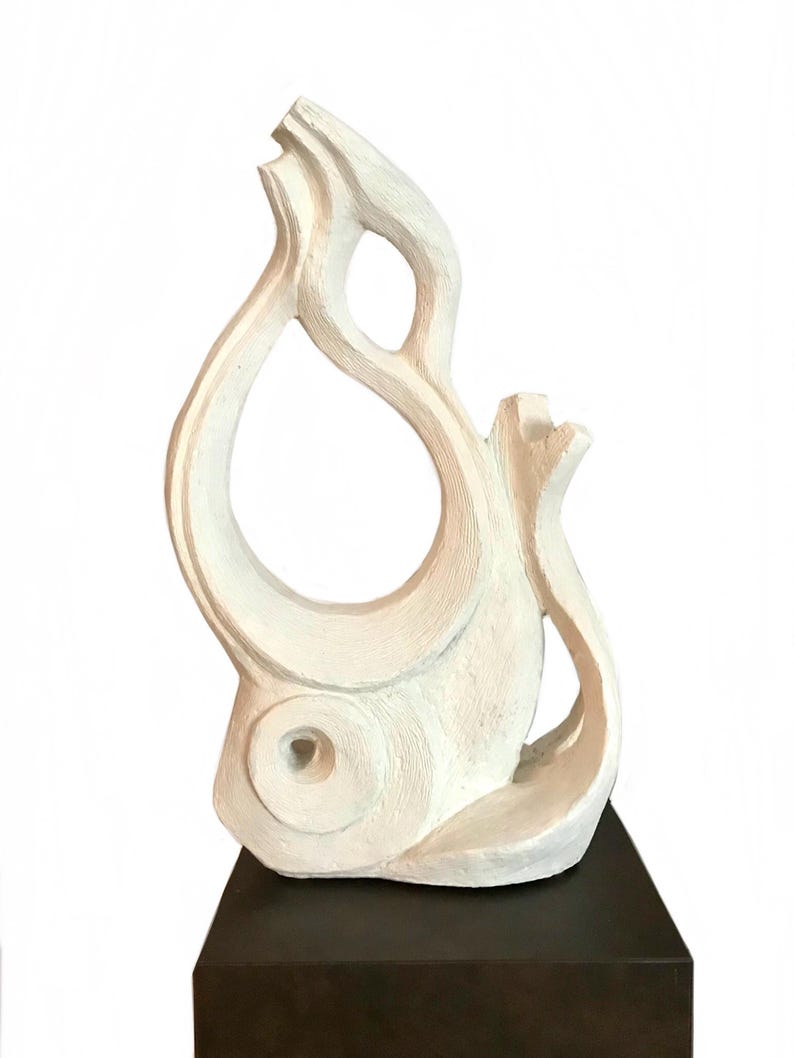 Pode incluir: Uma escultura abstrata em tom esbranqui&ccedil;ado com uma superf&iacute;cie texturizada e linhas esculpidas vis&iacute;veis. A pe&ccedil;a tem uma forma alta, curva e semelhante a uma chama, com um espa&ccedil;o central aberto e um recorte circular perto da base.