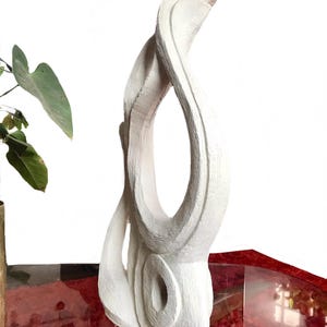 Pode incluir: Uma escultura abstrata alta de cor branca com uma superf&iacute;cie mate e texturizada. A pe&ccedil;a apresenta uma abertura vertical central e uma forma circular oca e arredondada na base, criando uma silhueta fluida com estrias vis&iacute;veis.