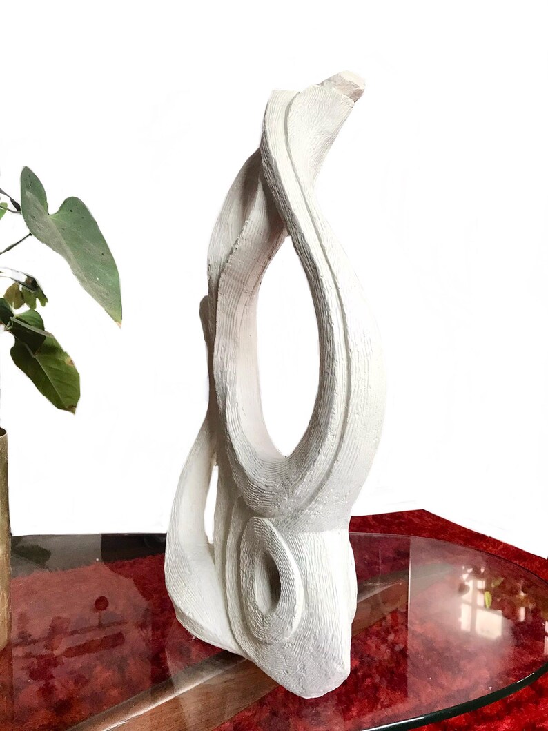Pode incluir: Uma escultura abstrata alta de cor branca com uma superf&iacute;cie mate e texturizada. A pe&ccedil;a apresenta uma abertura vertical central e uma forma circular oca e arredondada na base, criando uma silhueta fluida com estrias vis&iacute;veis.