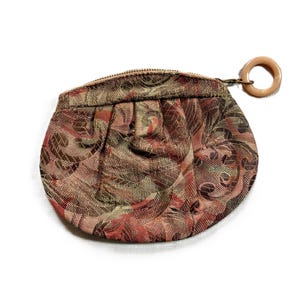 May include: A small, rounded coin purse featuring a woven tapestry fabric with a multi-colored floral and geometric pattern in shades of red, brown, and green. The purse has a top zipper closure finished with a circular, light beige plastic ring pull.