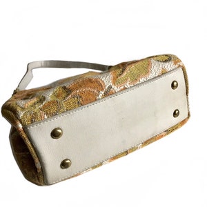 May include: A small, rectangular handbag with a white leather base and a fabric top featuring a floral pattern in shades of brown, yellow, and green. The bag has a single strap and gold-coloured metal accents.
