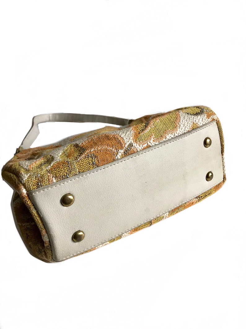 May include: A small, rectangular handbag with a white leather base and a fabric top featuring a floral pattern in shades of brown, yellow, and green. The bag has a single strap and gold-coloured metal accents.
