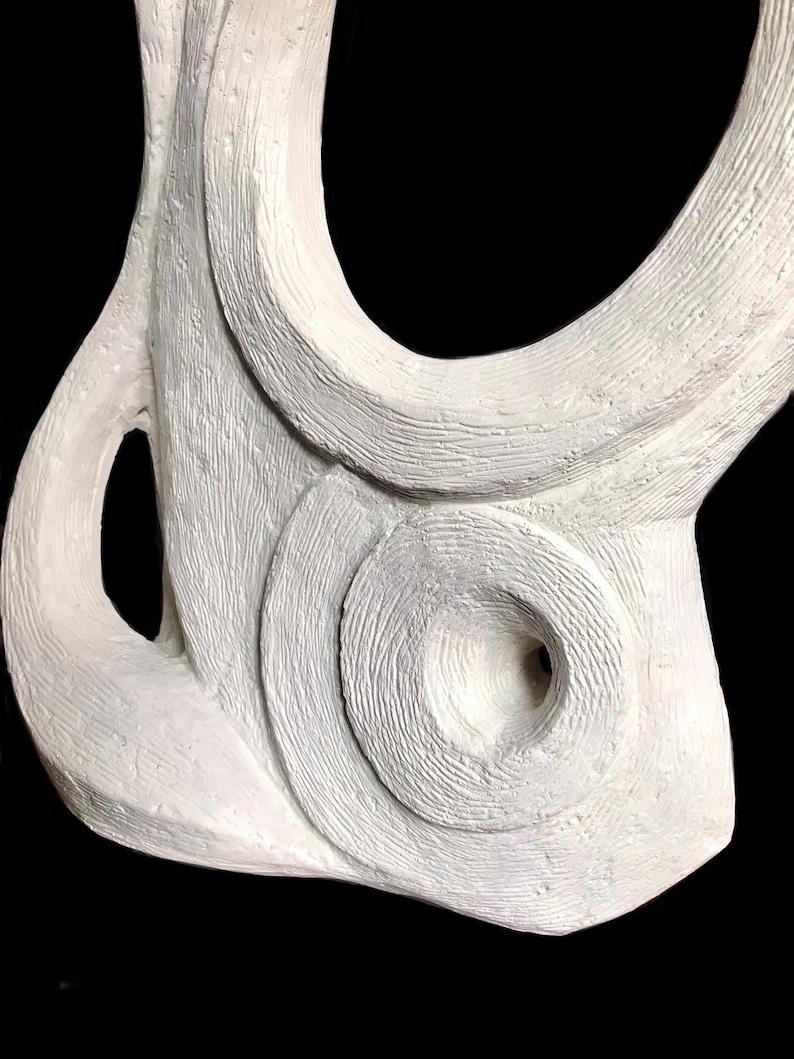 Pode incluir: Uma escultura abstrata branca com uma superf&iacute;cie texturizada com sulcos e linhas vis&iacute;veis. O objeto tem uma forma org&acirc;nica curva com uma reentr&acirc;ncia circular proeminente no centro e uma abertura no lado esquerdo.