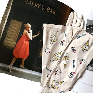 May include: A pair of white cotton gloves with a whimsical design featuring yellow umbrellas, keys, and perfume bottles. The gloves are lying on top of a magazine with the words "Harry's Bar" on the cover.