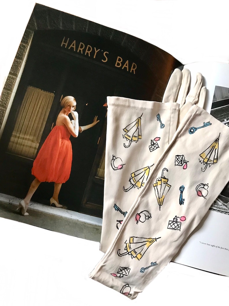 May include: A pair of white cotton gloves with a whimsical design featuring yellow umbrellas, keys, and perfume bottles. The gloves are lying on top of a magazine with the words "Harry's Bar" on the cover.