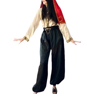 Peut inclure: Un haut crème à manches longues avec des bordures dorées et une ceinture noire avec une chaîne dorée. La tenue comprend un pantalon noir ample et une coiffe rouge avec des accents dorés. Des chaussures noires à bout ouvert complètent l'ensemble.