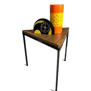 May include: A mid-century modern style side table with a black metal frame and a triangular wooden top. On the table are a black and yellow clock and a tall orange and yellow vase with a circular pattern.