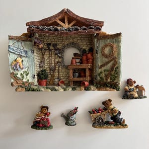 May include: A miniature garden shed diorama with a stone wall interior, gardening tools, and terracotta pots. The shed features a sign reading GARDENERS LEARN BY TROWEL AND ERROR. Four separate bear figurines are included: one with apples, a watering can, a bear with a wheelbarrow, and a bear with a seed bag.