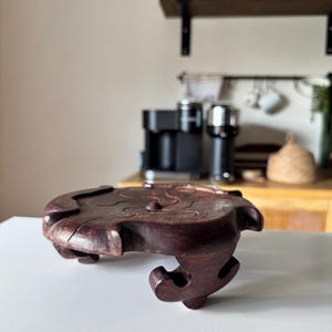 May include: A dark brown wooden display stand with a circular top featuring a carved spiral pattern and four curved, ornate legs. The stand has a smooth, polished finish and is designed to elevate objects, showcasing a sturdy and detailed construction.