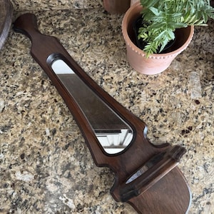 May include: A dark brown wooden wall-mounted mirror with a shelf. The mirror is oval-shaped and set within a decorative frame. The shelf is located at the bottom of the mirror. The wood has a rich, polished finish.