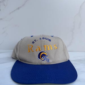 May include: A tan and blue St. Louis Rams baseball cap. The cap features the team name in gold embroidery, along with the team logo of a ram's head. The brim is a solid blue color.