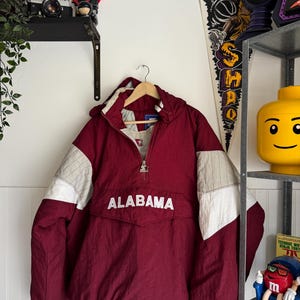 May include: A maroon and white Starter brand pullover jacket featuring a hood, a half-zip front closure with a metallic logo zipper pull, and the word ALABAMA embroidered in white across the center front pocket flap.