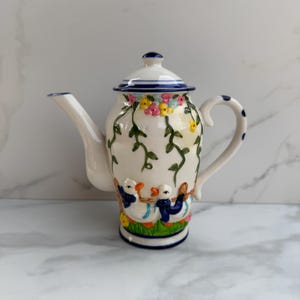 May include: A white ceramic teapot with a lid, spout, and handle. The teapot is decorated with a floral design, green vines, and images of ducks. The handle has blue polka dots. The base has a green and blue border.