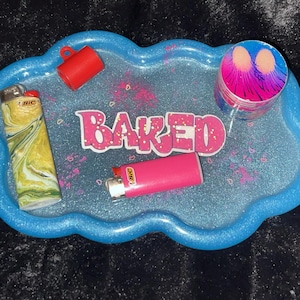 May include: A blue, cloud-shaped tray with the word "BAKED" in pink letters. The tray holds a red cup, a pink lighter, a yellow and green lighter, and a colorful grinder. The tray has a glittery finish.