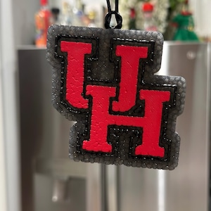 Puede incluir: Un ambientador con el logotipo UH rojo y negro, colgado de una cuerda negra. Las letras UH son rojas con un contorno negro, sobre un fondo gris texturizado. El ambientador es probablemente para un coche o una habitación.