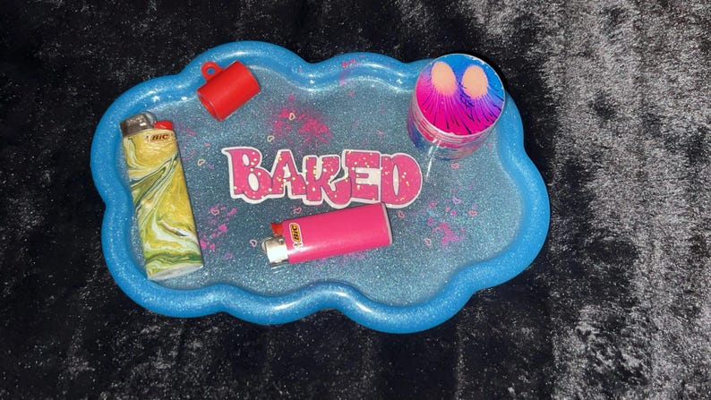 May include: A blue, cloud-shaped tray with the word "BAKED" in pink letters. The tray holds a red cup, a pink lighter, a yellow and green lighter, and a colorful grinder. The tray has a glittery finish.