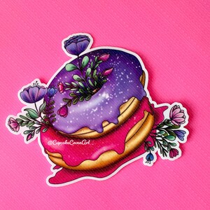 May include: A purple and pink donut with a galaxy glaze and white sprinkles, topped with a cluster of purple and pink flowers. The donut is dripping with pink glaze and the text "@CupcakeCovenArt" is visible on the side.