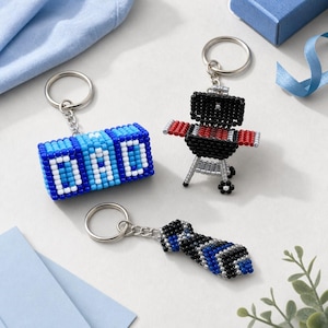May include: Three beaded keychains featuring a rectangular block with the word DAD in white letters on a blue background, a black and red charcoal grill with silver legs, and a necktie pattern in shades of blue, black, and silver beads.