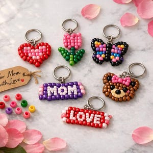 May include: A collection of handmade keychains in various designs, including a heart, a tulip, a butterfly, a bear, and the words "MOM" and "LOVE". The keychains are made of colorful beads and are arranged on a marble surface with rose petals.