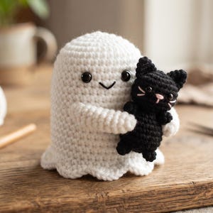 Op de afbeelding: Een wit gehaakt spookfiguurtje met zwarte kraaloogjes en een gestikte mond, dat een kleine zwarte gehaakte kat vasthoudt met een roze gestikte neus en snorharen. Het spookje heeft een ronde kop en een geschulpte onderrand.