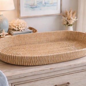 Puede incluir: Una cesta ovalada tejida con asas de cuerda. La cesta es de color marrón claro con un patrón texturizado. Las asas son de cuerda trenzada. La cesta está sobre una superficie de madera, lo que sugiere un artículo decorativo o de almacenamiento.