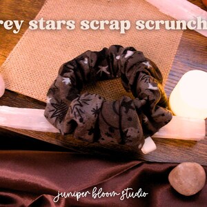 May include: A grey scrunchie with a star and leaf pattern, resting on a burlap surface. The text "grey stars scrap scrunchie" is at the top. The image also includes crystals and the text "juniper bloom studio".