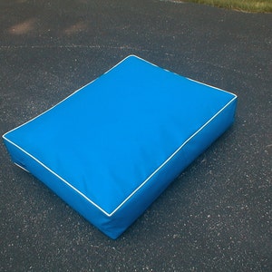 May include: A rectangular, bright blue cushion with white piping around the edges. The cushion is placed on a dark surface, and appears to be made of a smooth, durable material. This cushion could be used as a pet bed or a decorative pillow.