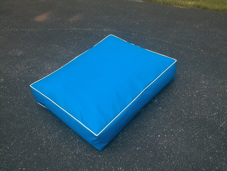 May include: A rectangular, bright blue cushion with white piping around the edges. The cushion is placed on a dark surface, and appears to be made of a smooth, durable material. This cushion could be used as a pet bed or a decorative pillow.