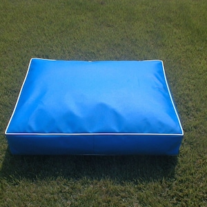 May include: A rectangular, bright blue pet bed with white piping around the edges. The bed is made of a smooth, durable fabric and is designed for comfort. The bed is placed on a green grassy surface.