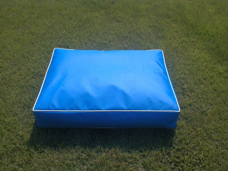 May include: A rectangular, bright blue pet bed with white piping around the edges. The bed is made of a smooth, durable fabric and is designed for comfort. The bed is placed on a green grassy surface.