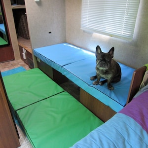 May include: A small, black and brown dog sits on a blue and green bed in a camper. The bed is made of two separate pieces, one blue and one green. The dog is looking at the camera.