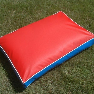 May include: A rectangular pet bed with a red top, blue sides, and white piping. The bed is placed on green grass. The dimensions are approximately 30 inches long, 20 inches wide, and 4 inches thick.