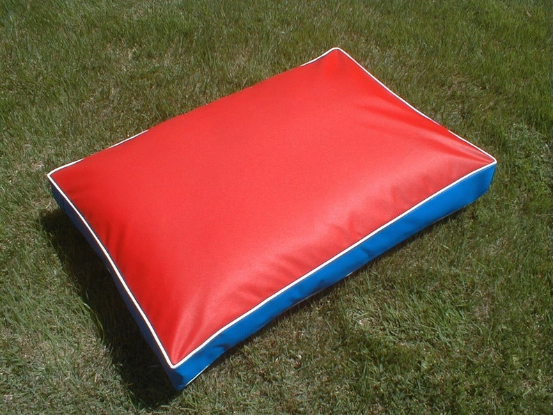 May include: A rectangular pet bed with a red top, blue sides, and white piping. The bed is placed on green grass. The dimensions are approximately 30 inches long, 20 inches wide, and 4 inches thick.