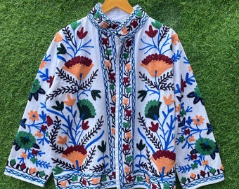 May include: White jacket with colorful floral embroidery. The jacket features orange, green, blue, and red flower designs. The jacket is hanging on a wooden hanger against a green grass background.