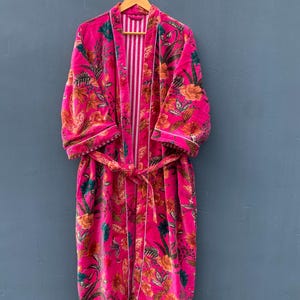 May include: A vibrant pink floral robe with a white trim and a matching belt. The robe features a colorful floral pattern with orange, green, and yellow accents. The interior has a vertical striped pattern.