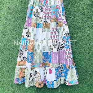 Op de afbeelding: Lange patchwork rok met diverse bloemen- en geometrische patronen in blauw, paars, oranje en groen. De rok heeft een gefranjerde zoom en een elastische tailleband. De rok hangt aan een zwarte hanger tegen een groene achtergrond.