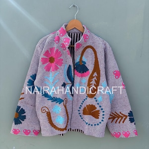 May include: A lavender jacket with a herringbone pattern and floral embroidery in pink, blue, and brown. The jacket has a striped lining and the text "NAIRAHANDICRAFT". It is hanging on a wooden hanger.