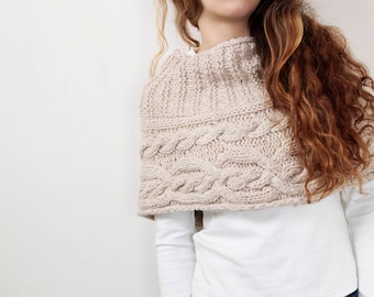 May include: A woman wearing a beige knitted cable knit cowl neck sweater with a white long-sleeved shirt and blue jeans.