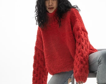 May include: A person wearing a chunky, bright red turtleneck sweater with openwork detailing on the sleeves. They are wearing light blue jeans and black high-top sneakers. The person is sitting on a gray stool.