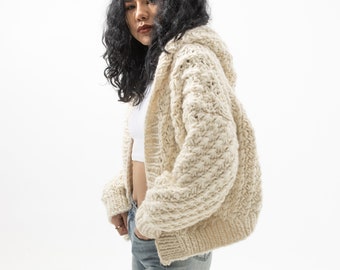 Puede incluir: Un cárdigan con capucha de punto grueso de color crema. El suéter abierto presenta un patrón de puntadas texturizadas y mangas largas. La modelo lleva vaqueros azules de lavado claro y una camiseta blanca.