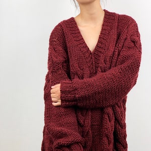 May include: A burgundy cable-knit sweater with a deep V-neck. The oversized jumper has a loose fit and long sleeves. The knitwear features a textured cable design on the front and sleeves. The model is wearing blue jeans.