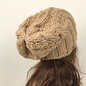 May include: A beige knitted beanie with a cable knit pattern. The beanie is slouchy and has a loose fit.