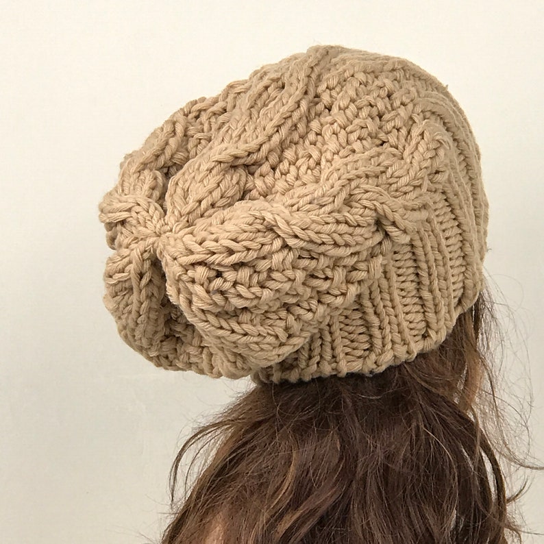 May include: A beige knitted beanie with a cable knit pattern. The beanie is slouchy and has a loose fit.